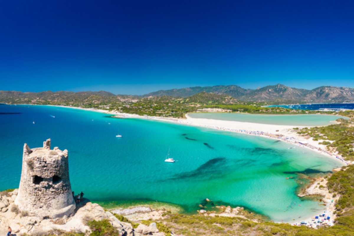 Vista panoramica di Spiaggia di Simius al tramonto, sabbia bianca e mare turchese, cielo dai colori caldi, presenza di bagnanti e ombrelloni